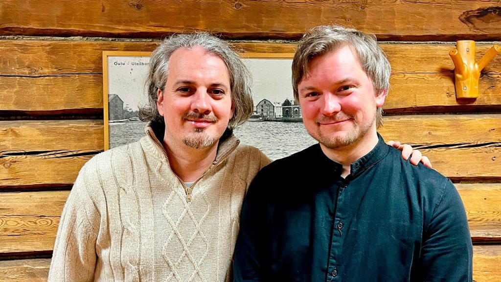 Two men pose for the camera in front of a wooden wall.