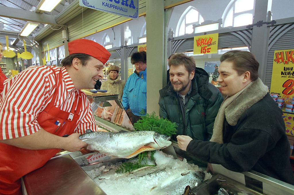 Kaksi miestä kalatiskillä kauppahallissa, kauppias tiskin toisella puolella.