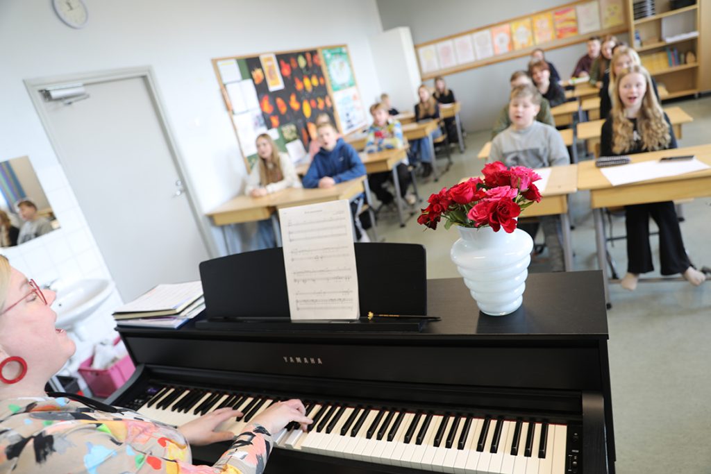 Musiikinopettaja laulaa ja soittaa pianoa, taustalla oppilaita pulpeteissaan.