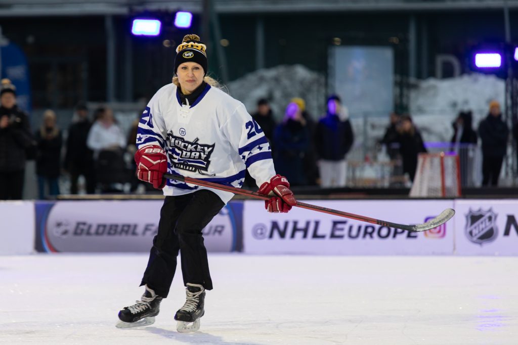 Nainen luistelee jääkiekkomaila kädessään ja pipo päässään valkoisessa pelipaidassa.