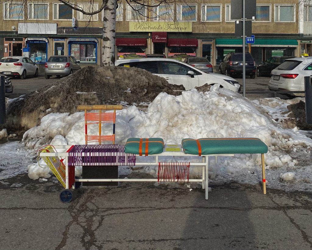 Erilaisilla vaatekappaleilla ja verhoiluilla koristeltu penkki katukuvassa.