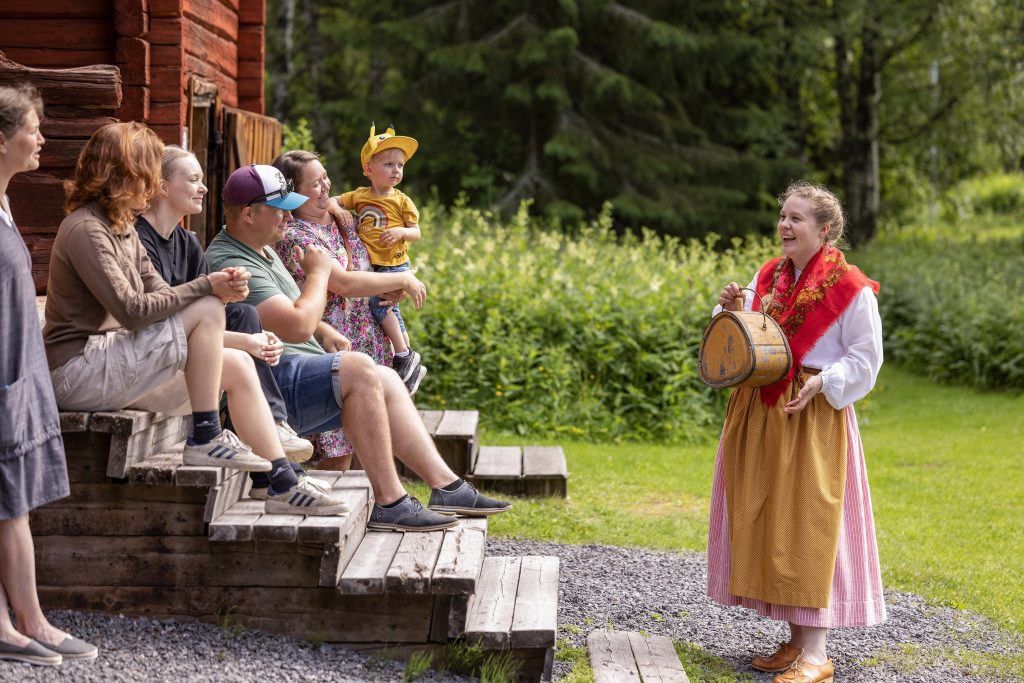 Kansallispuvussa oleva nainen puhuu aitan portailla istuvalle ryhmälle.