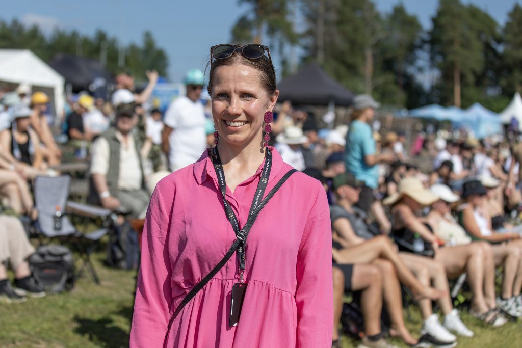 Nainen vaaleanpunaisessa mekossa hymyilee auringossa, taustalla raviyleisöä.