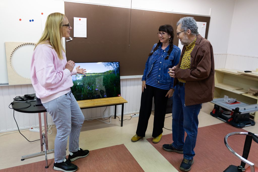 Kaksi naista ja yksi mies seisovat ison tv-näytön ympärillä.