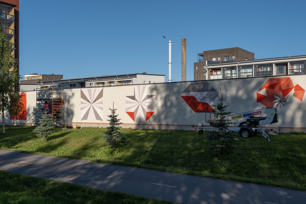 Seinämaalaus tiilimuurissa, taustalla kerrostaloja ja tehtaiden piippuja.