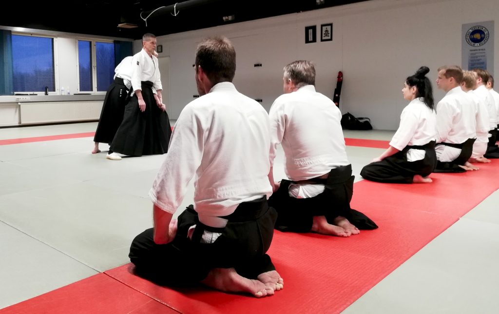 Ihmisiä polvillaan rivissä aikidoharjoituksissa, opettaja edessä.