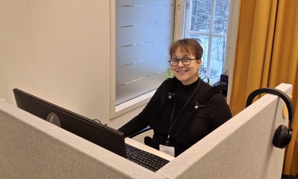 Silmälasipäinen, lyhythiuksinen nainen hymyilee tietokoneen näytön takana.