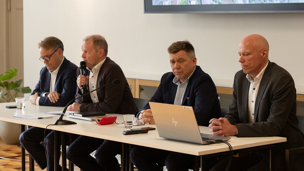 Neljä miestä pöydän ääressä tiedotustilaisuudessa, yksi puhuu mikrofoniin.