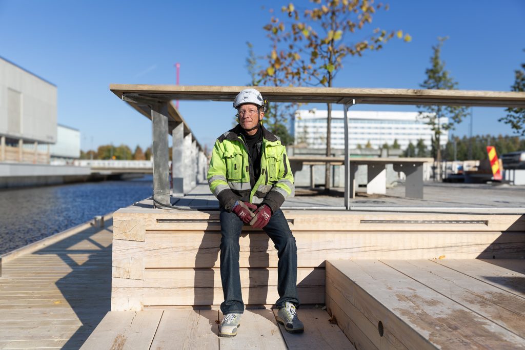 Työmaavaatteisiin pukeutunut mies istuu hirsilaiturilla.