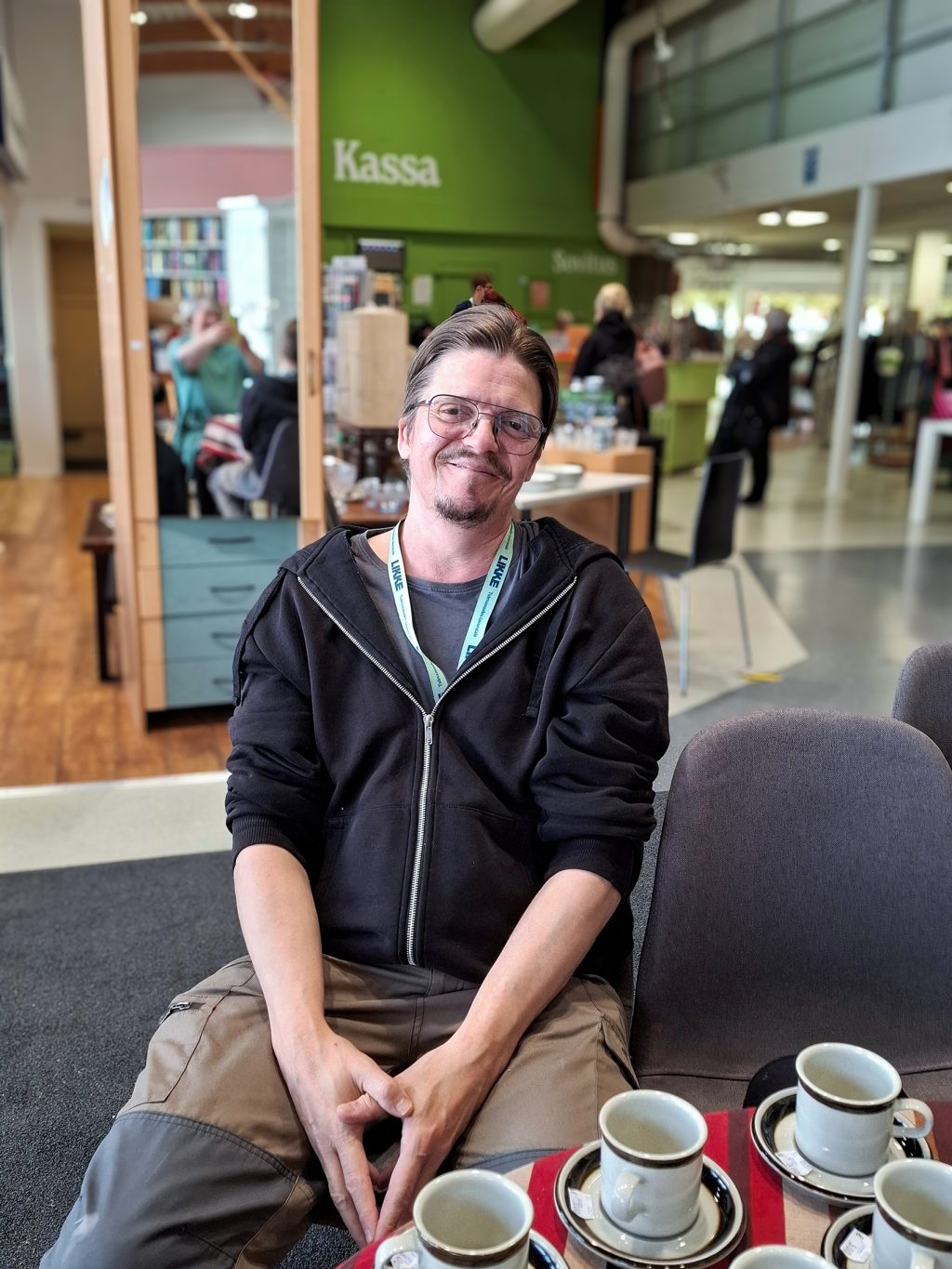 Silmälasipäinen mies istuu kierrätyskeskuksen myymälässä tuolilla, etualalla pöydällä kahvikuppeja.