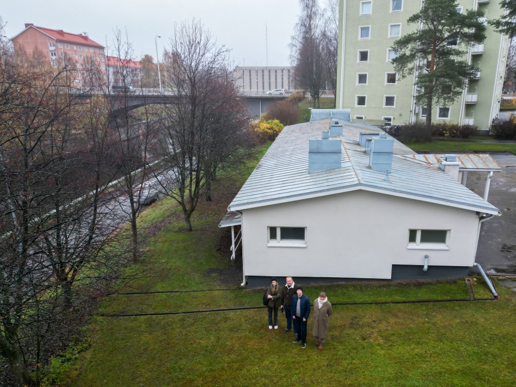 Ilmakuva saunasta, jonka edustalla seisoo neljä opiskelijaa.