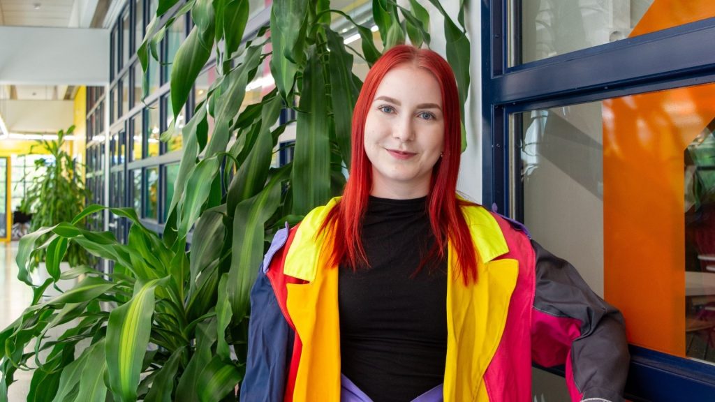 Punahiuksinen nuori nainen värikkäässä takissa puolilähikuvassa käytävällä viherkasvin edessä.