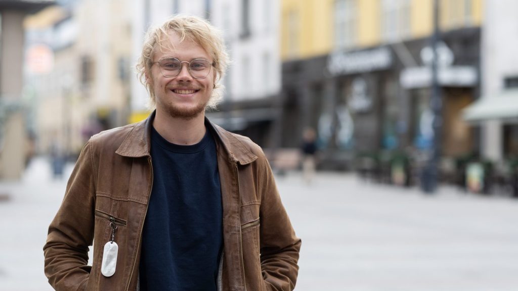 Silmälasipäinen vaalea nuori mies ruskeassa takissa puolilähikuvassa kävelykatuaukiolla.