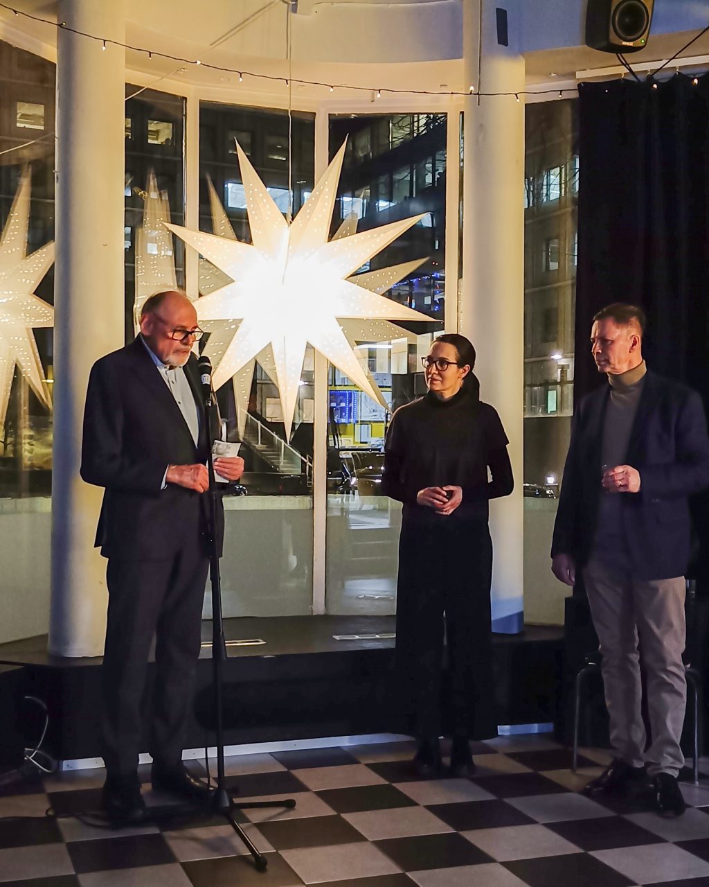 Mies pitää seison puhetta, vierellä kuuntelemassa nainen ja mies. Taustalla ikkunassa suuri joulutähti.