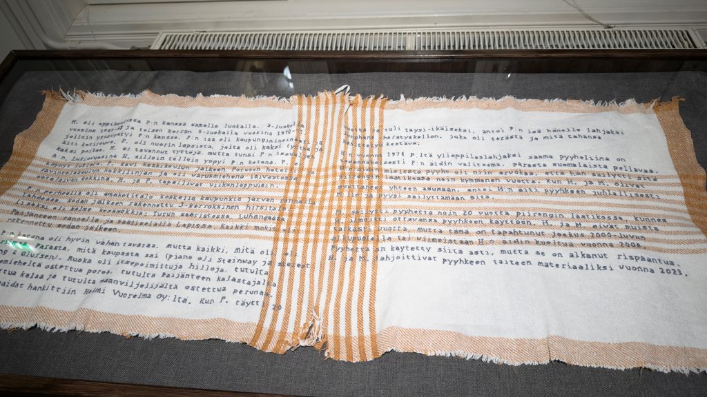 Taideteos, jossa vanhaan pyyheliinaan kirjailtuna tekstiä.