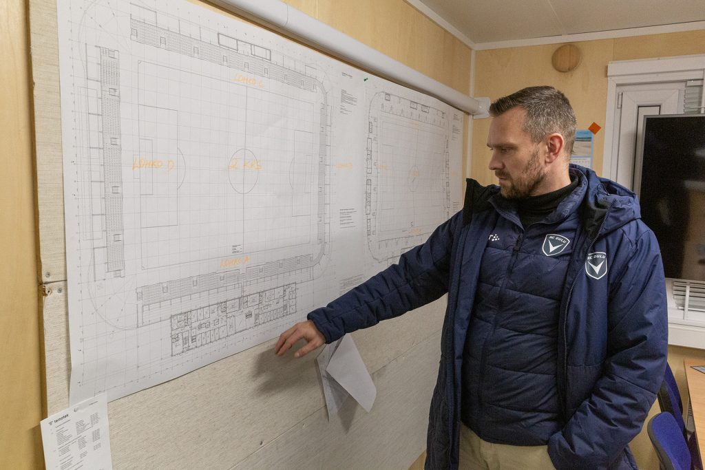 Sinitakkinen mies osoittaa kädellä seinällä olevaa suurta rakennuspiirustusta.