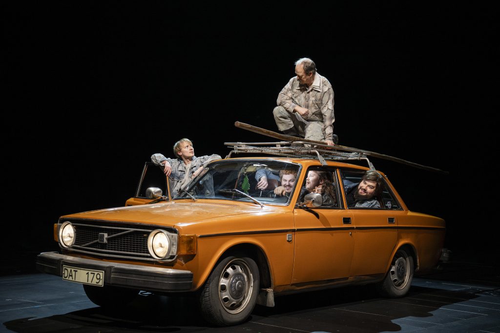 Näyttelijöitä lavalla oranssissa autossa, yksi auton katolla.