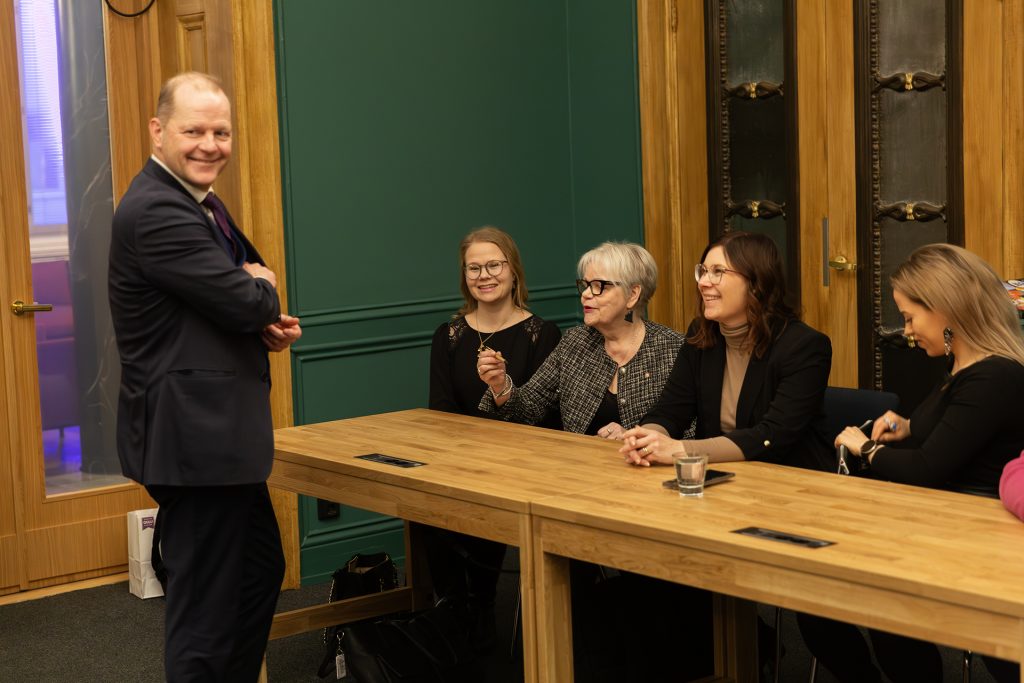 Kaupunginvaltuutettuja pöydän ääressä, edessä seisoo hymyilevä kaupunginjohtaja joka kaivaa povitaskuaan.