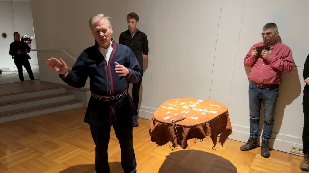 Mies nahasta tehdyn rummun vieressä, taustalla näyttelyn yleisöä.