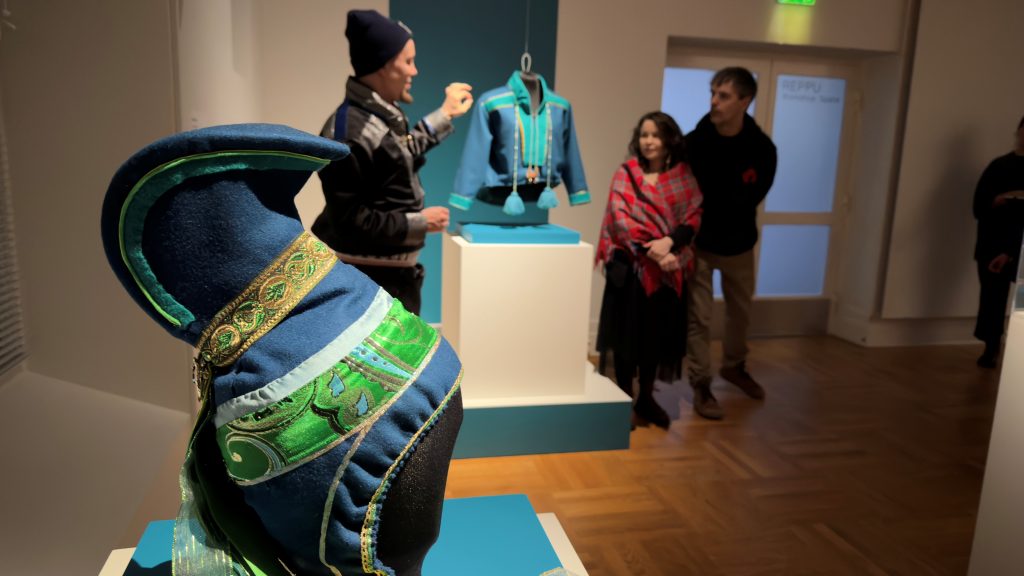 Etualalla vitriinin päällä sininen saamelaishattu, taustalla kaksi miestä ja nainen saamenpuvun ympärillä.