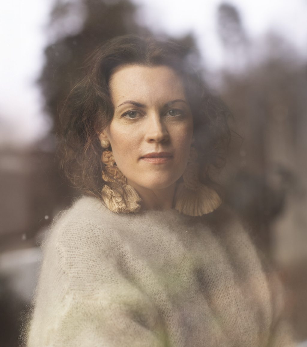 Tummahiuksinen nainen vaaleassa villapaidassa utuisessa puolilähikuvassa.