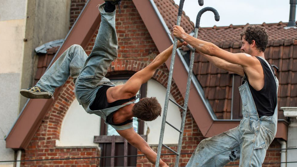 Kaksi miesakrobaattia kiipeilee köysillä rakennuksen edessä.