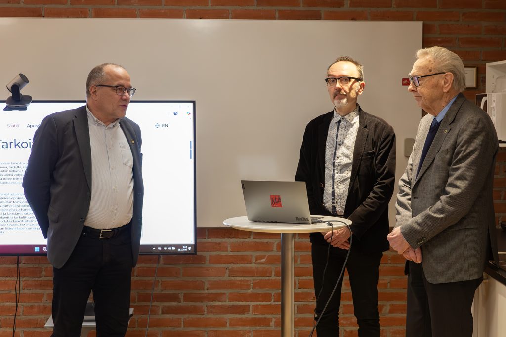 Kolme miestä seisoo, pyöreällä pöydällä keskellä kannettava tietokone.