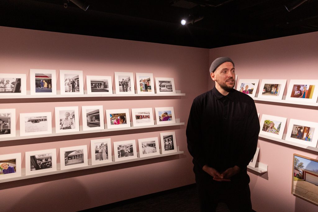 Mies seisoo näyttelytilassa, taustalla pieniä valokuvateoksia vaaleanpunaisella seinällä.