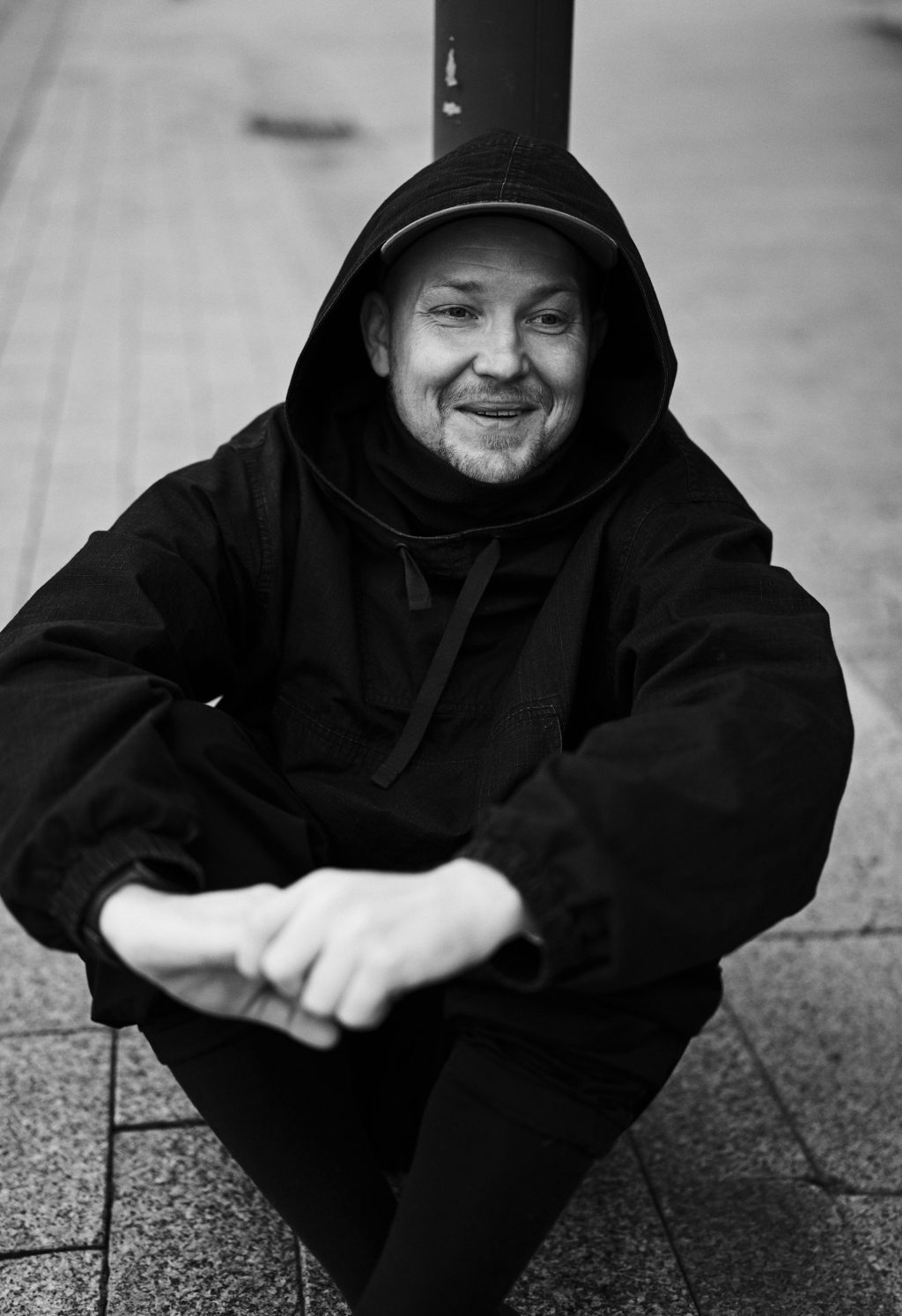 Hymyilevä huppupäinen mies istuu jalat koukussa mustavalkoisessa kuvassa.