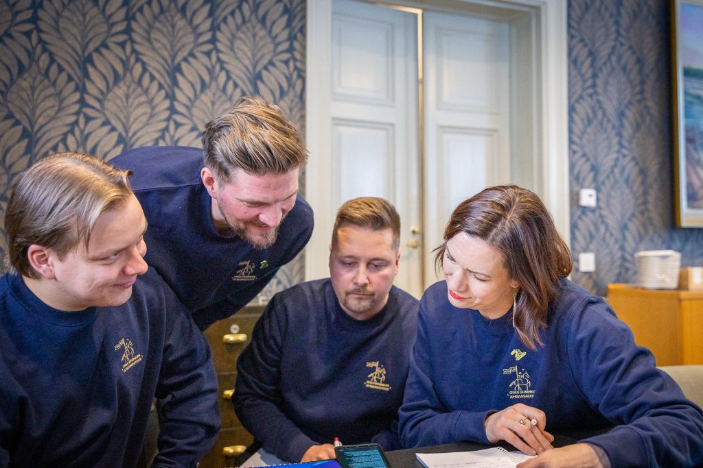 Kolme miesta ja yksi nainen kumartuneena tutkimaan kännykän näyttöä.