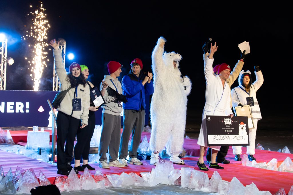Joukko ihmisiä tuulettaa punaisella matolla, yksi pukeutunut jääkarhuasuun, yhdellä suuri shekki kädessä.