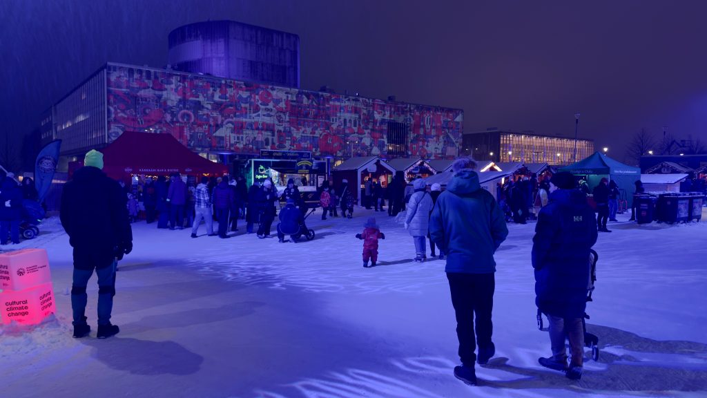 Ihmisiä talvisella torilla sinisessä valaistuksessa, taustalla teatteritalo jonka seinään heijastettu värikäs kuvio.