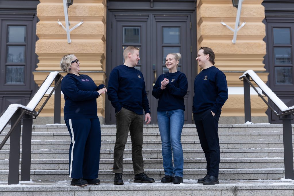 Kaksi naista ja kaksi miestä samanlaisissa sinisissä college-puseroissa seisovat kaupungintalon portailla ja nauravat.