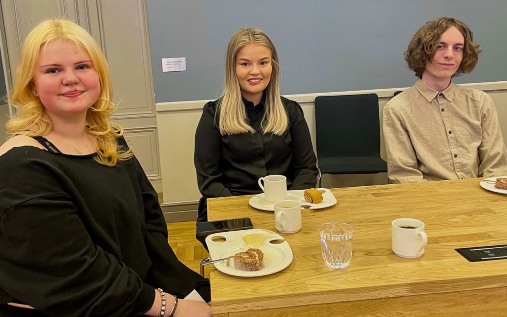 Kaksi nuorta naista ja nuori mies kahvipöydässä.