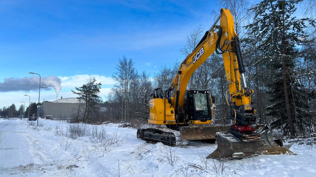 Kaivinkone lumisen tien ja metsän välissä, taustalla palloiluhalli.