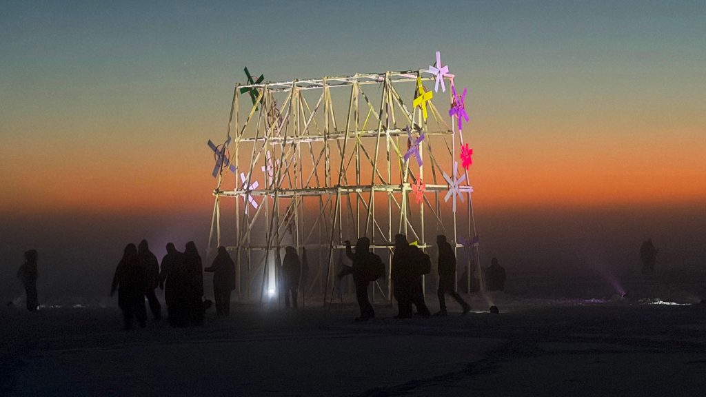 Ihmisiä taide-installaation ympärillä meren jäällä auringon laskiessa.