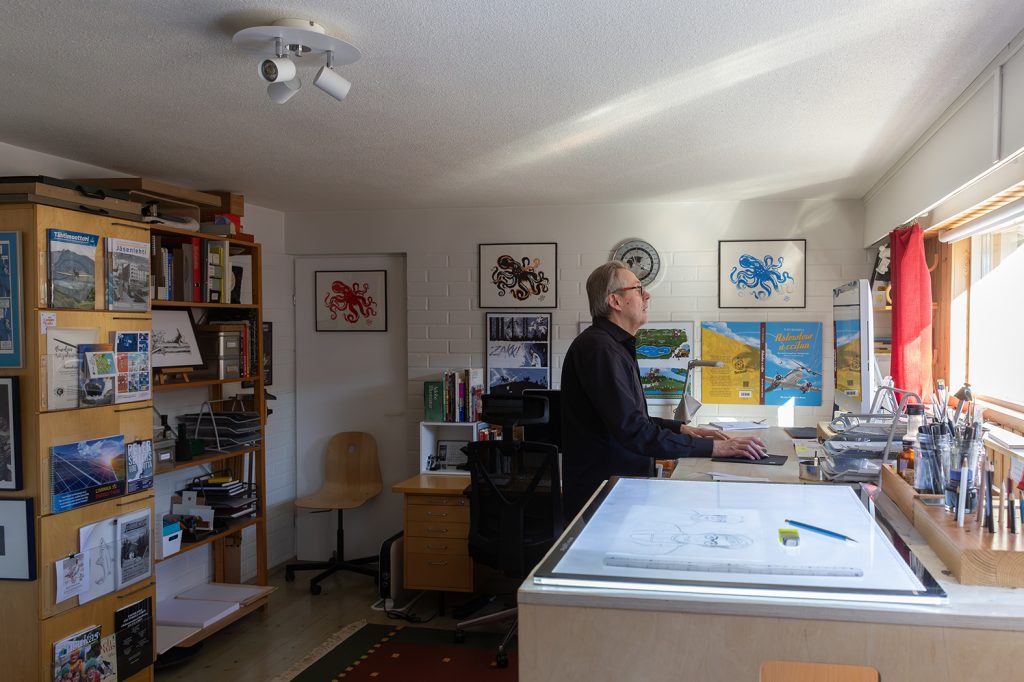 Mies työhuoneessa tietokoneen äärellä, seinillä paljon kehystettyjä piirroksia ja julisteita.