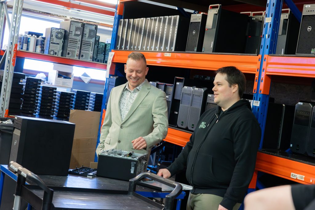 Kaksi miestä pöydän ääressä, taustalla hyllyssä tietokoneita.