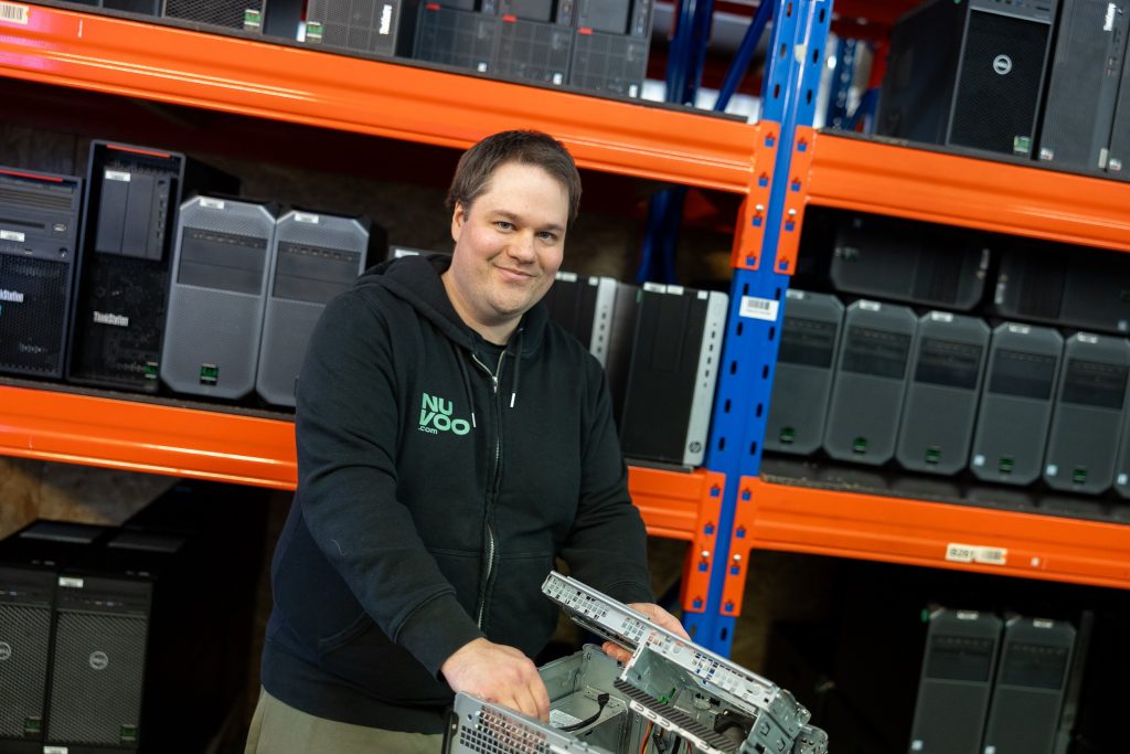 Nuorehko mies mustassa hupparissa tietokoneen runko käsissään, taustalla hyllyssä rivi pöytätietokoneiden keskusyksikköjä.