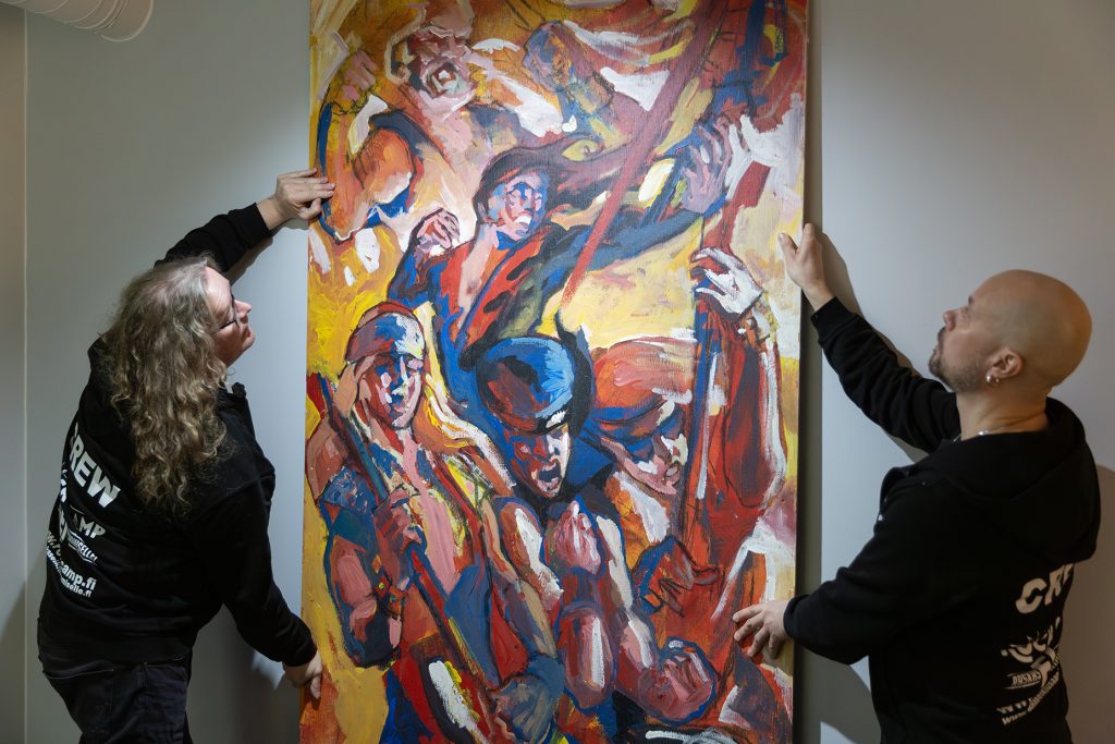 Kaksi miestä nostaa suurta, värikästä maalausta seinälle.