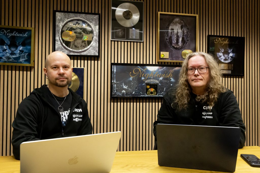 Kaksi mustiin pukeutunutta miestä, pöydällä kannettavat tietokoneet, taustalla seinällä kultalevyjä kehyksissä.