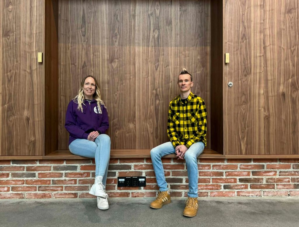 Nainen ja mies istuvat käytävän syvennyksessä, taustalla puuseinä.