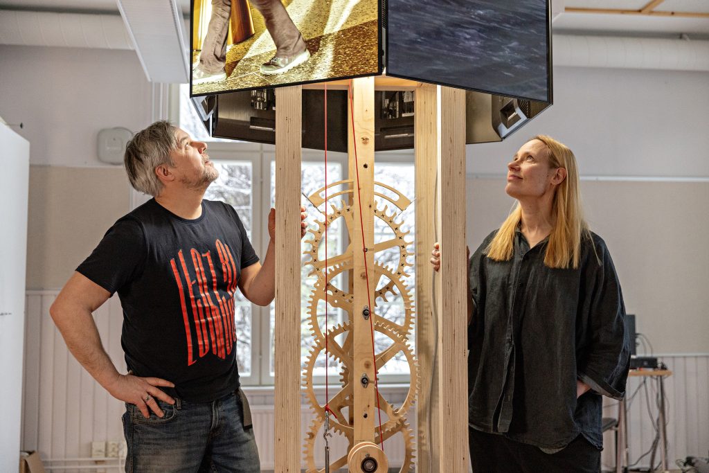 Mies ja nainen katselevat välissään olevaa suurta puista kelloa.