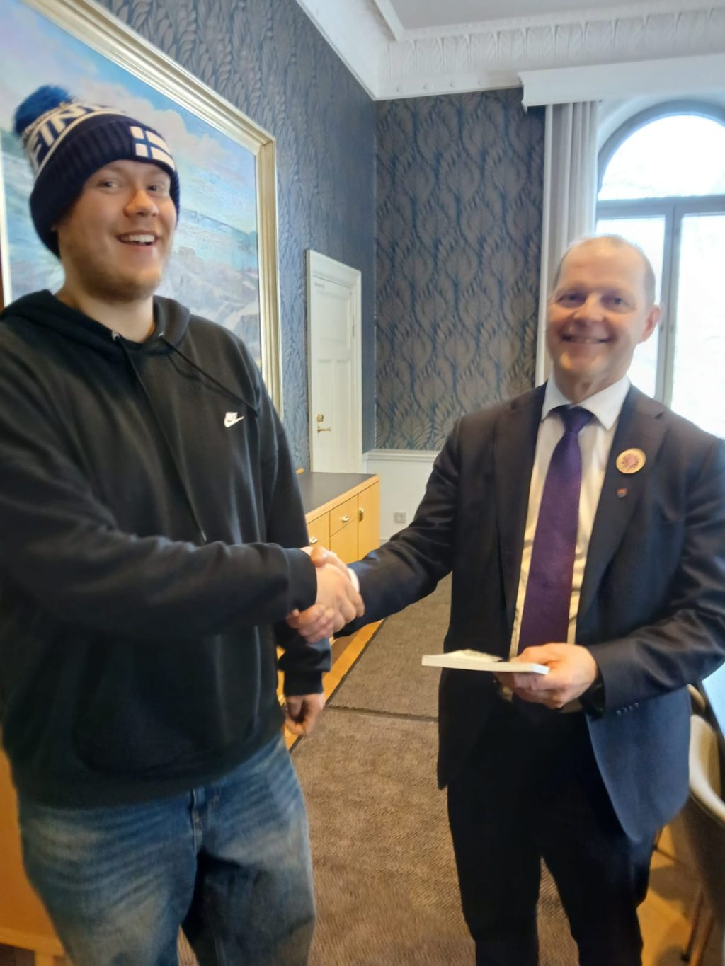 Two men shaking hands, one wearing a beanie and a hoodie and one wearing a suit