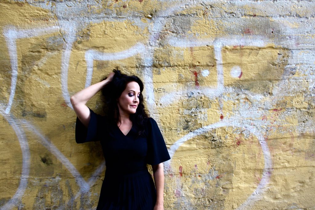 Tummaan mekkoon pukeutunut nainen poseeraa sivulle katsoen keltaisen, spraymaalatun seinän edessä.