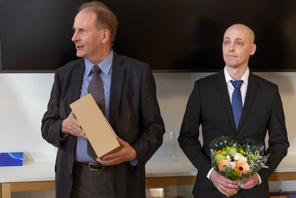 Kaksi miestä puvuissa, toisella palkintolaatikko ja toisella kukat käsissään.