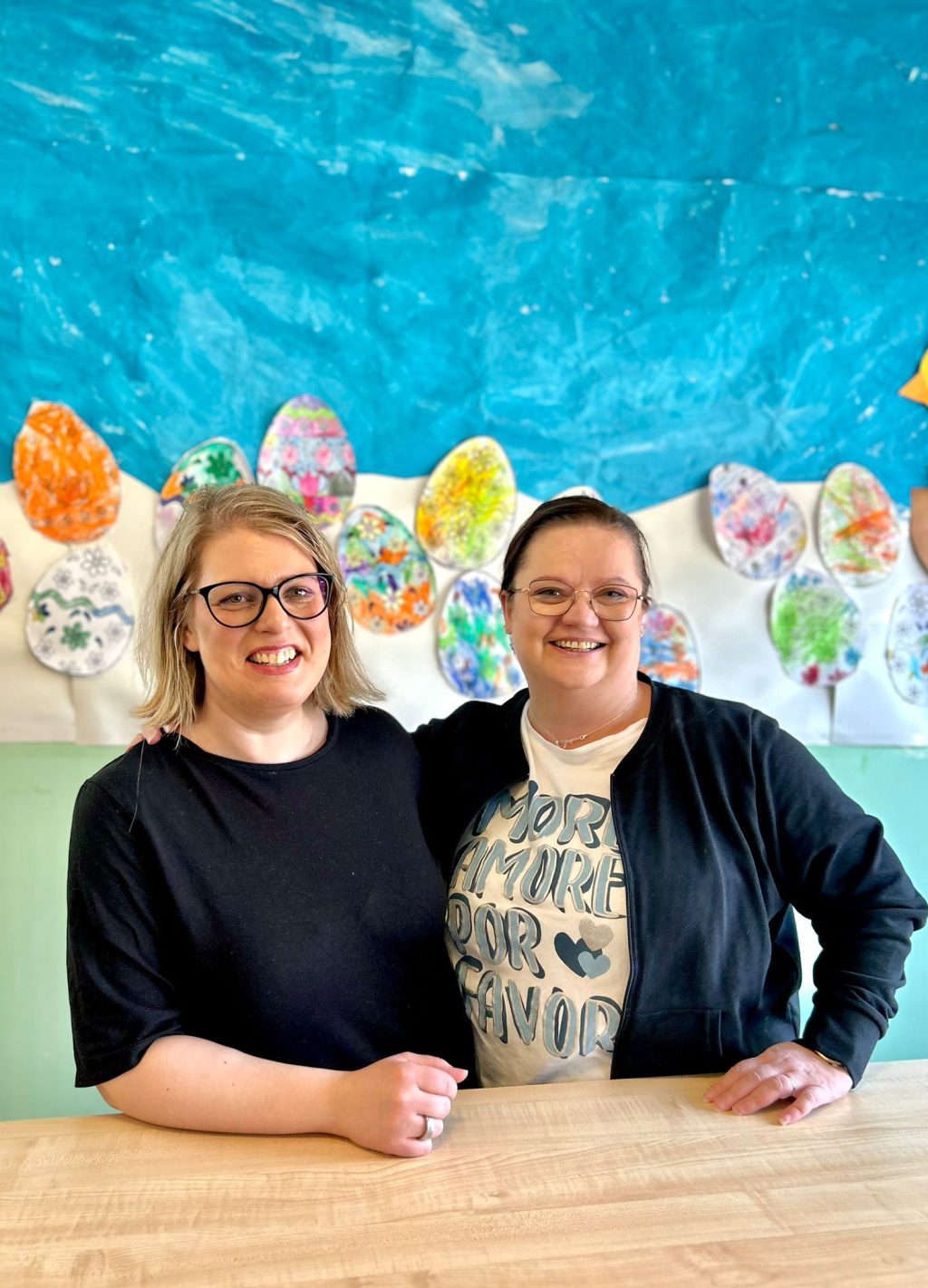 Kaksi naista poseeraa, seinällä taustalla lasten piirroksia.