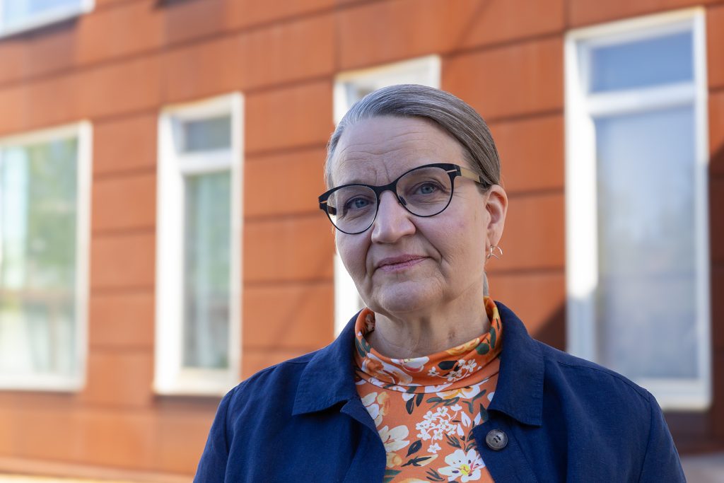 Silmälasipäinen nainen puolilähikuvassa, taustalla talon punaruskea seinä.
