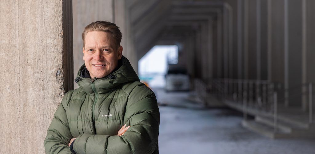 Mies vihreässä toppatakissa poseeraa betonisen teollisuusrakennuksen edessä.