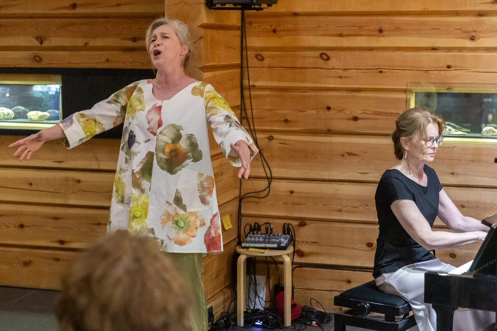 Vaaleatukkainen nainen laulaa hirsiseinän edessä, oikealla säestää silmälasipäinen nainen pianolla.